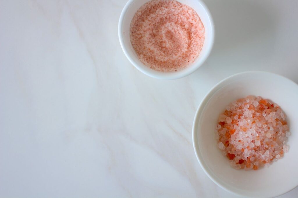 Two white ceramic bowls containing different textures of pink Himalayan salt on a marble surface.