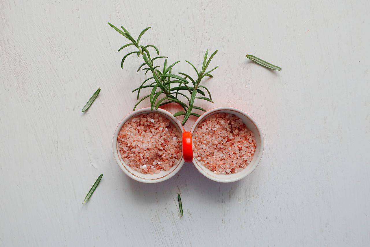 Top view of pink Himalayan salt and fresh rosemary in bowls on white table.