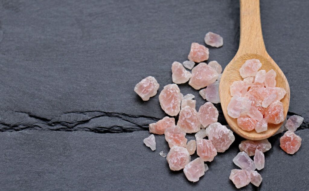 A wooden spoon with Himalayan pink salt crystals on a dark slate surface.